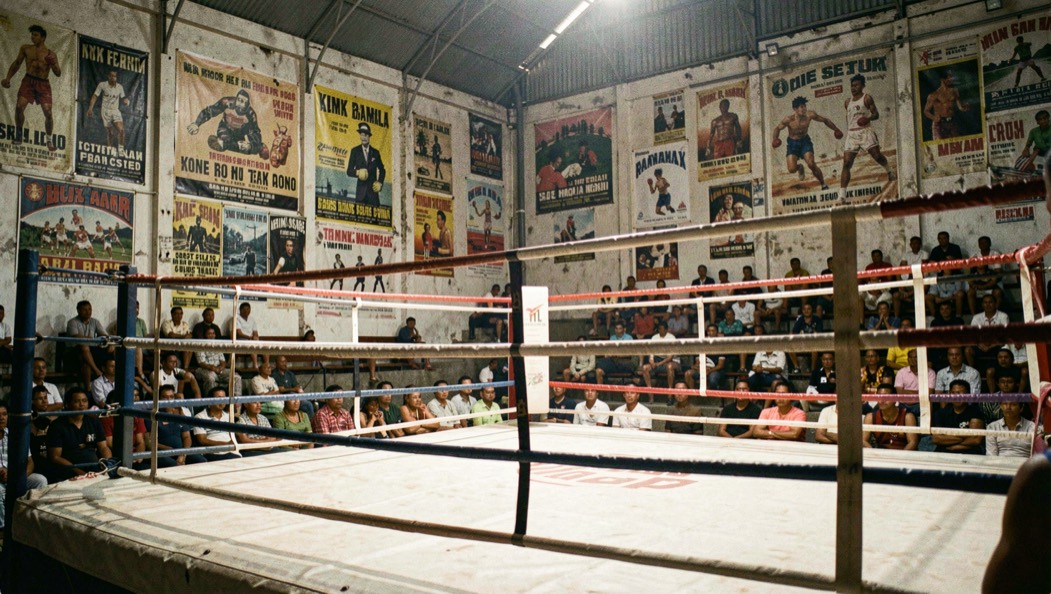 Grada de un estadio de boxeo en Bangkok con carteles antiguos en la pared y la lona del ring iluminada antes de la velada